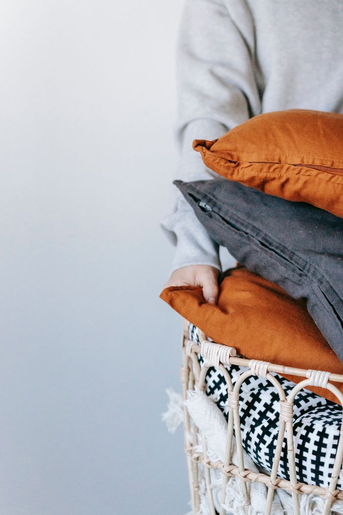 Person organizing a wicker basket with colorful throw pillows in a minimalist setting.